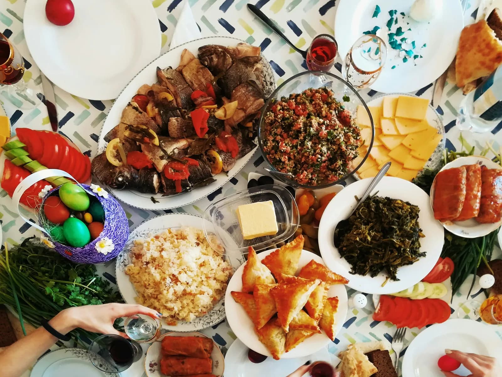 An overhead view of a crowded potluck table: a platter of whole roasted fish with peppers and lemon, a big bowl of tabbouleh, sliced cheese, golden hand pies, rice, greens, sliced tomatoes, and a basket of colored eggs, with hands reaching in for food and drinks.